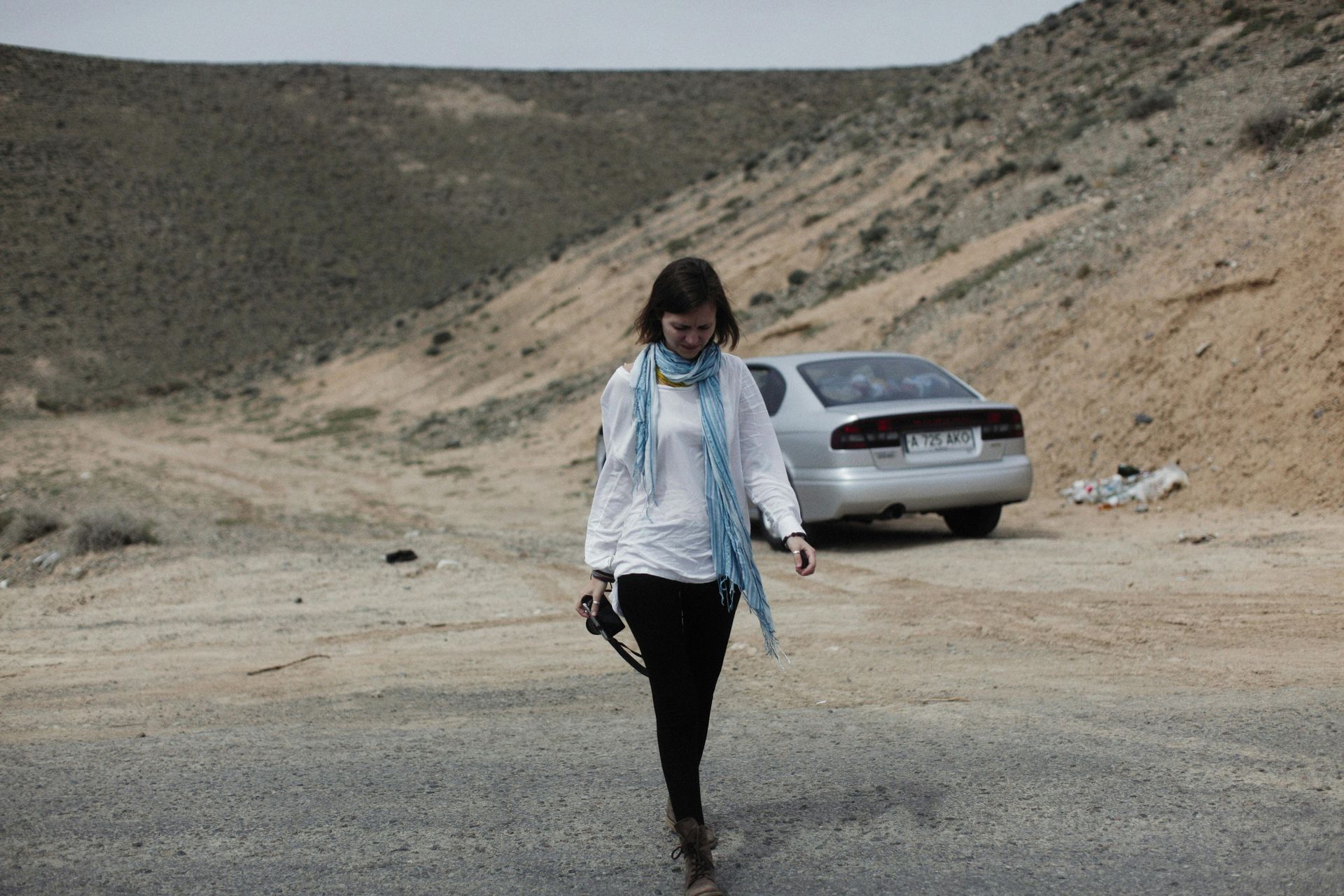 Woman walking on a dirt road near a parked silver car in a dry, rocky landscape