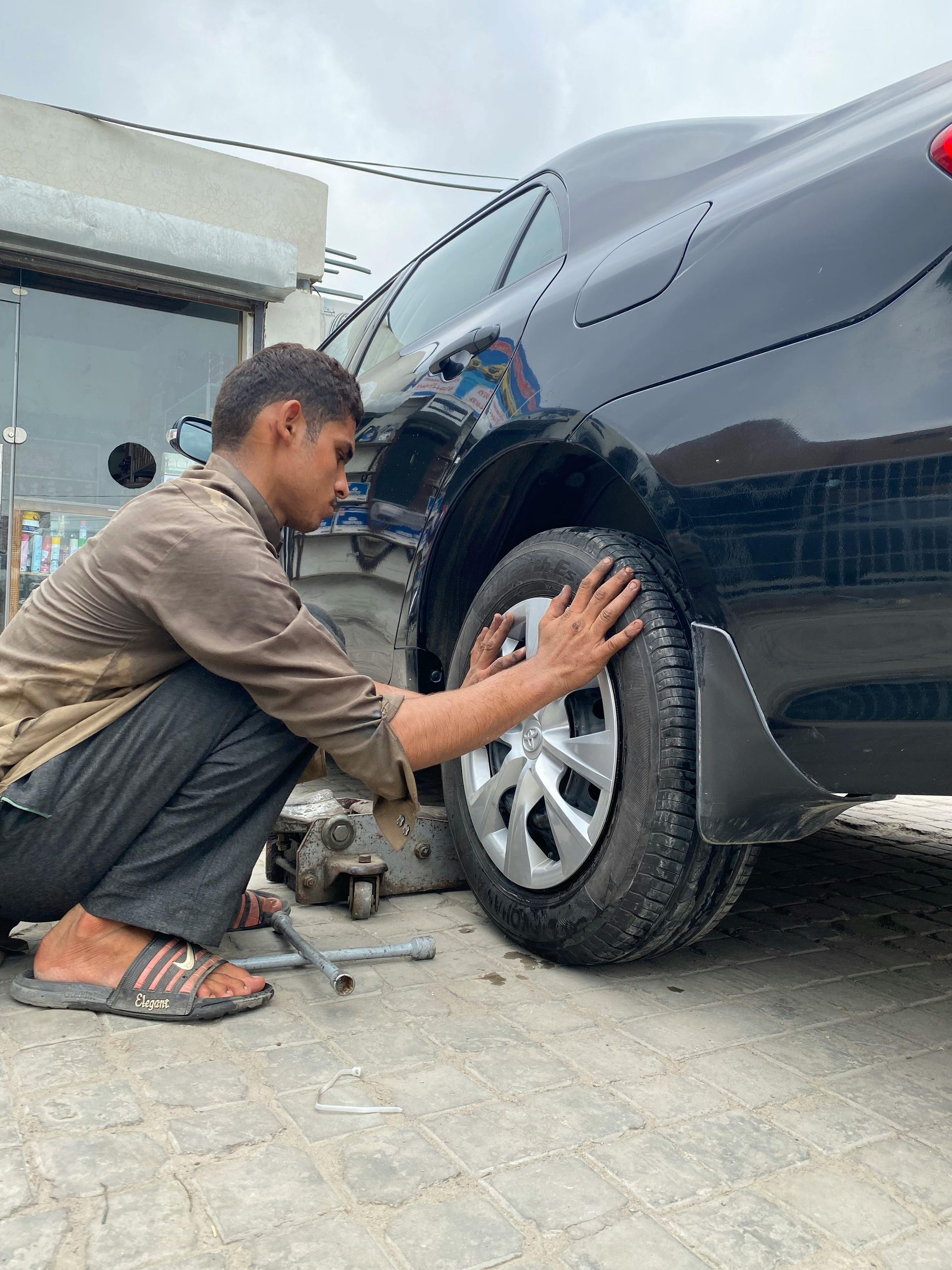 Person changing a car tire; kneeling beside black car, gray tires, and silver hubcap.