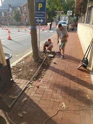 Workers repairing a sidewalk beside a street, with orange cones and a parked bike nearby.