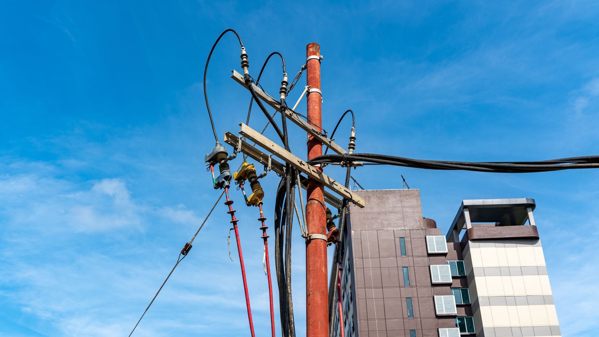 Utility pole with tangled cables against a blue sky, beside apartment buildings