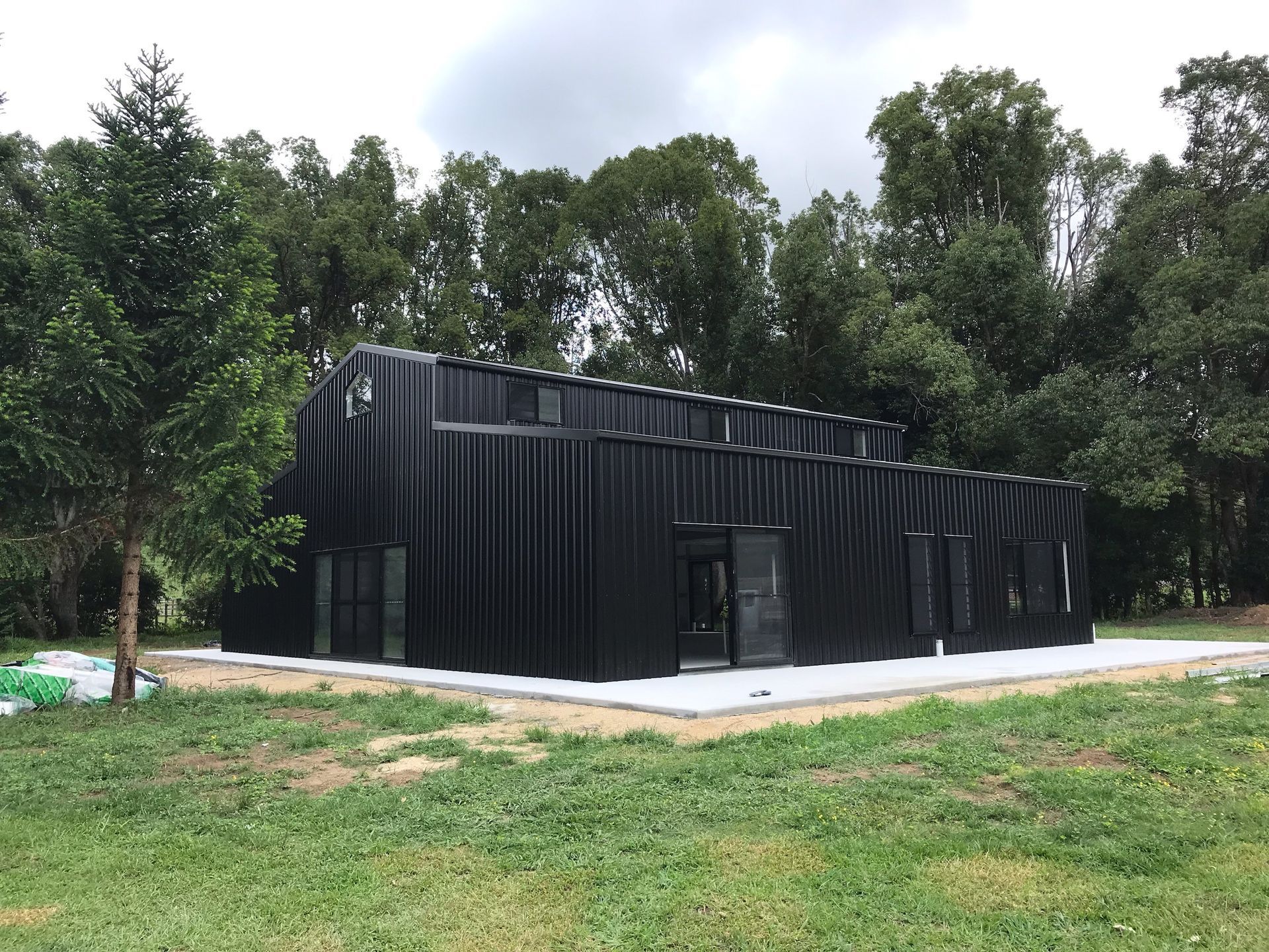 Black, modern building with a corrugated exterior, surrounded by trees and grass, on a concrete slab.— Northern Rivers Sheds in South Lismore, NSW