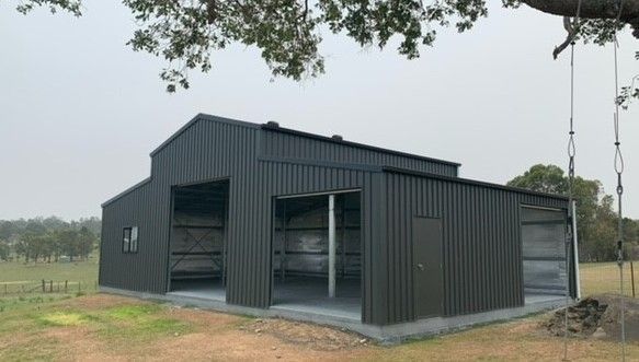 Dark grey metal barn structure with open bays, concrete floor, set on a green lawn under an overcast sky— Northern Rivers Sheds in Byron Bay, NSW