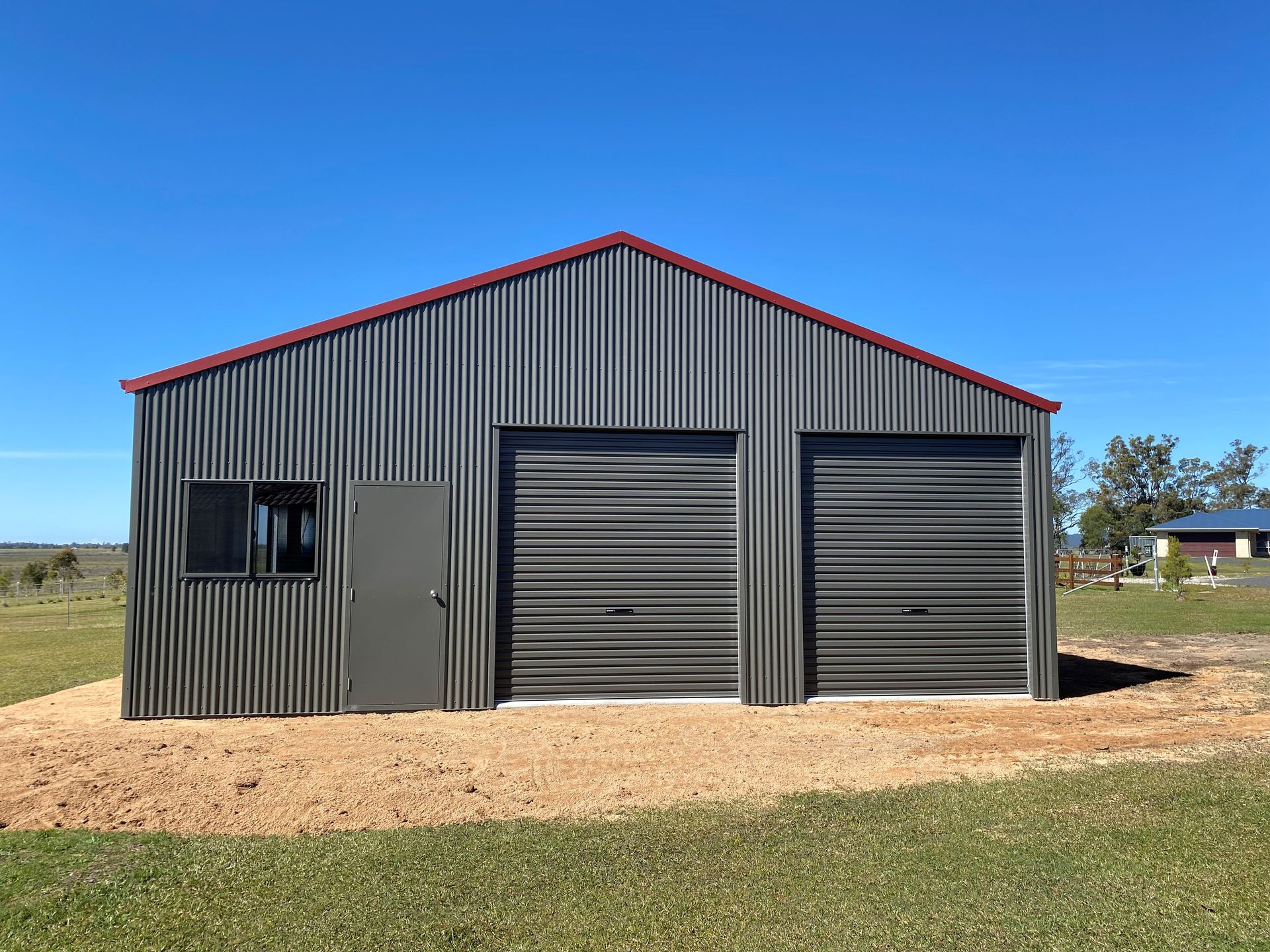 Steel gray corrugated shed with two garage doors, side door, and window; red trim, gravel base, blue sky— Northern Rivers Sheds in South Lismore, NSW