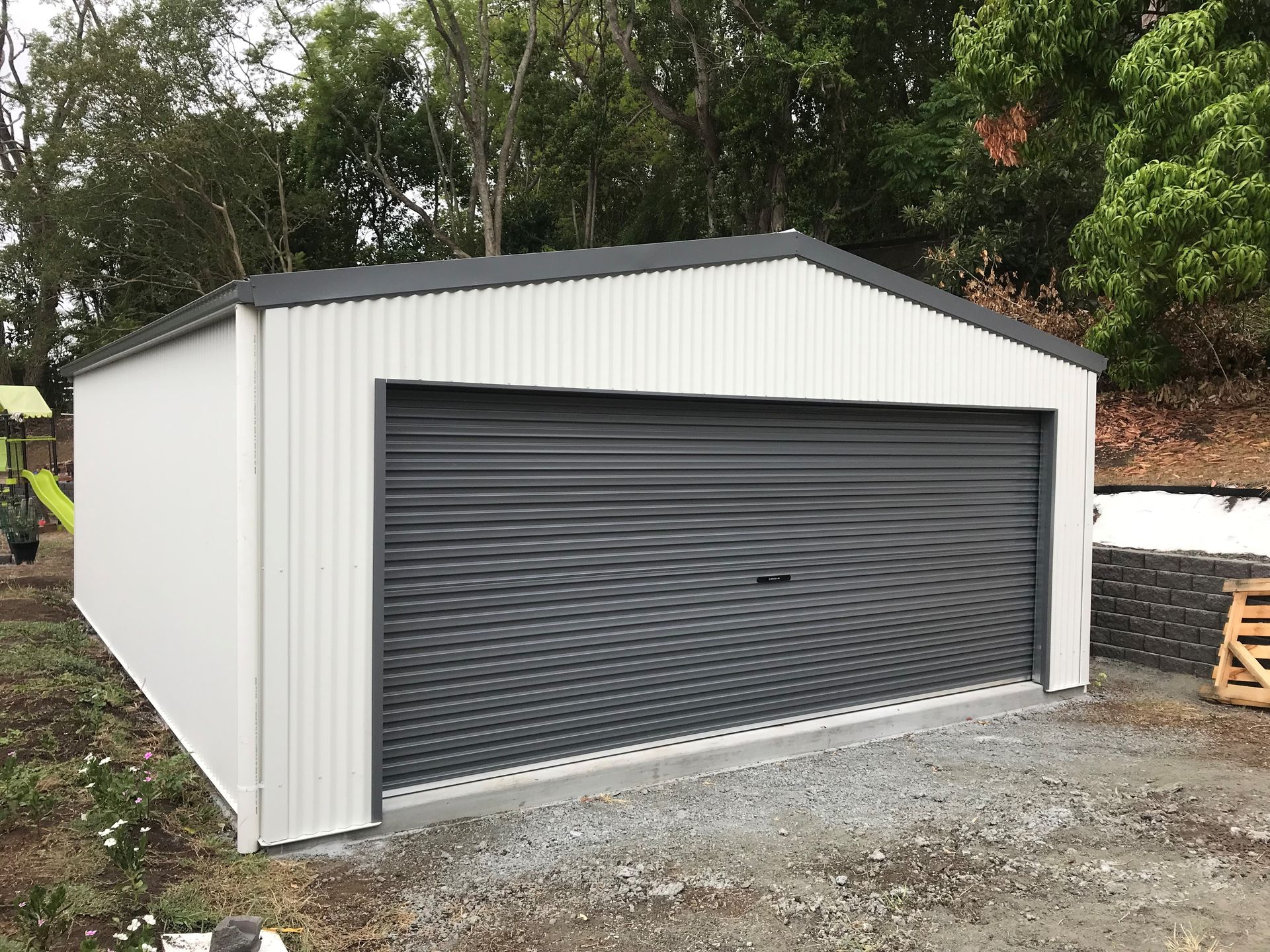 White garage with a gray roll-up door, on a gravel surface, against a green background— Northern Rivers Sheds in Byron Bay, NSW