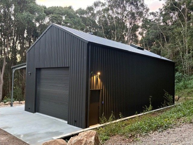 Black shed with a roller door and side door, concrete base, surrounded by trees— Northern Rivers Sheds in South Lismore, NSW