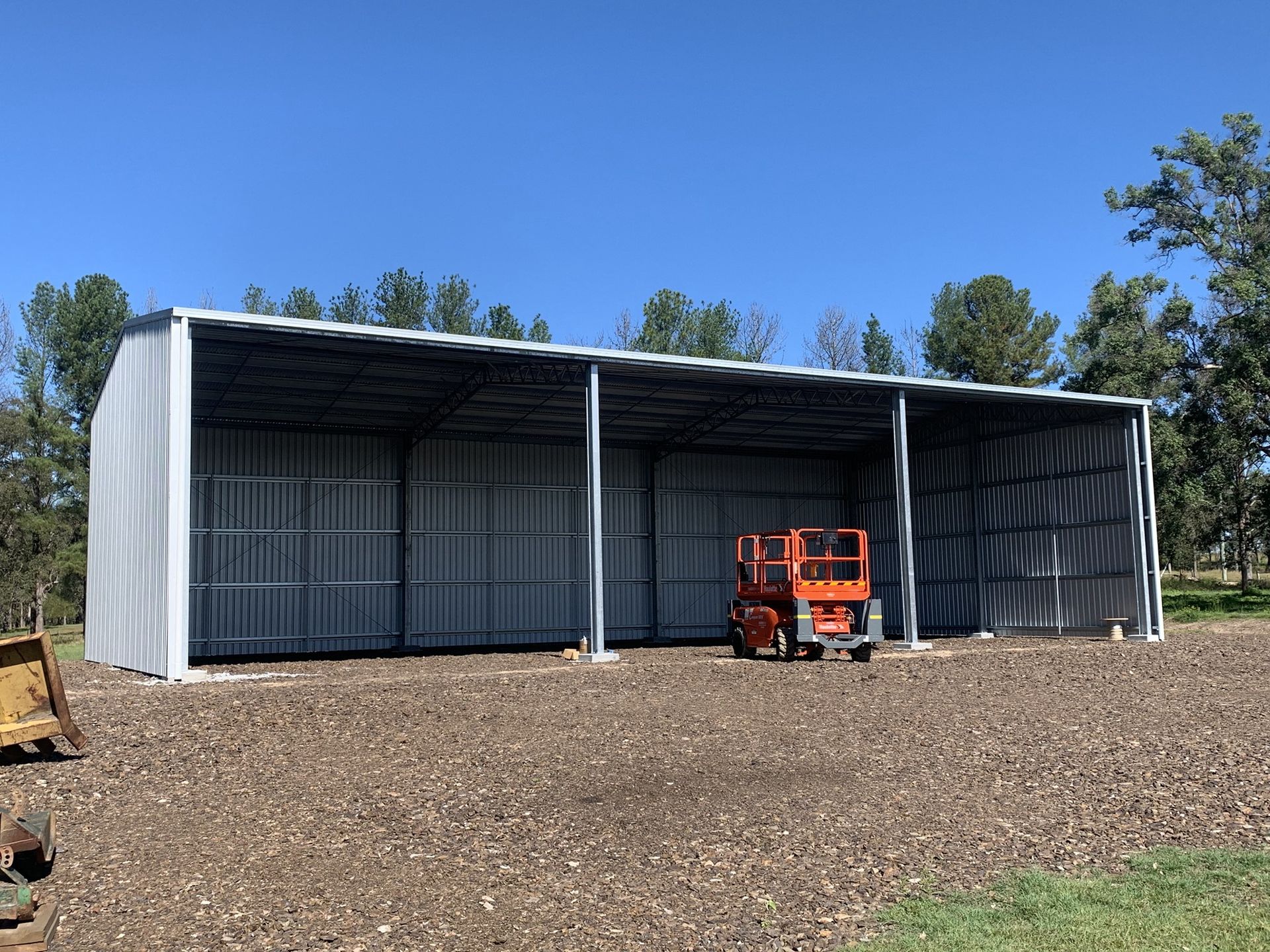 Metal open-sided agricultural shed with a lift inside, set against a blue sky with trees.— Northern Rivers Sheds in South Lismore, NSW