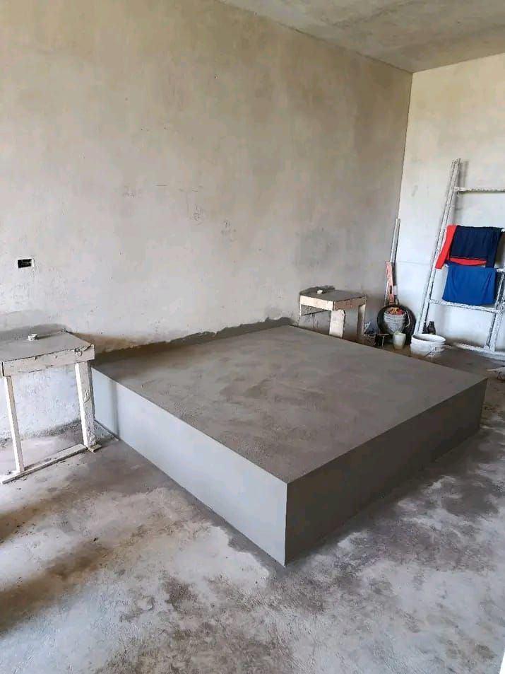 En una habitación con paredes sin terminar, cerca de una mesa blanca y una escalera, se alza una plataforma de hormigón de reciente construcción.
