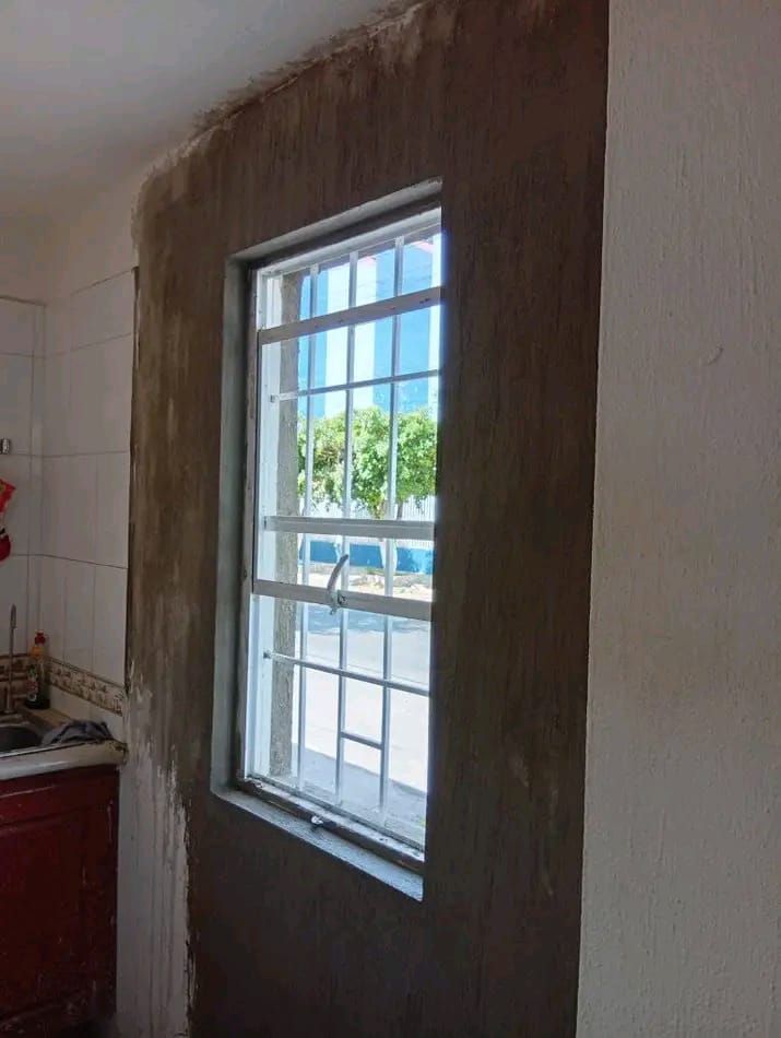 Una ventana en la pared de la cocina que muestra un parche de mortero marrón fresco alrededor de su marco, lo que evidencia las reparaciones en curso en la vivienda.