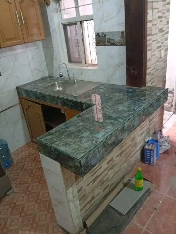 Una encimera de cocina con fregadero, con superficies que imitan el mármol verde y una base de azulejos, colocada contra una pared alicatada.