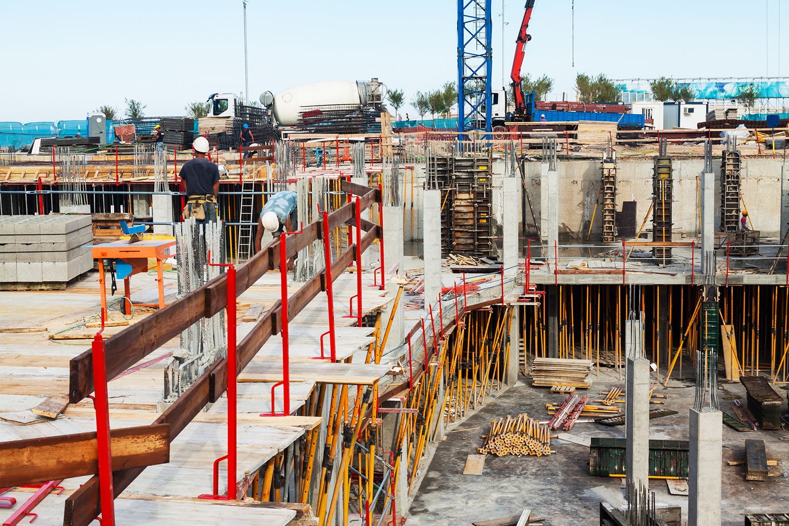 Obra en construcción con trabajadores, grúas, cimientos de hormigón y barandillas de seguridad de madera.