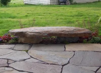 A large rock bench is sitting on a stone patio in front of a lush green field.