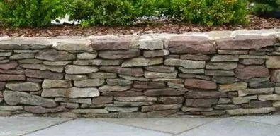 A stone wall is sitting on a sidewalk next to a bush.
