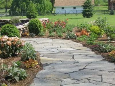 A stone walkway in a garden with flowers and a house in the background