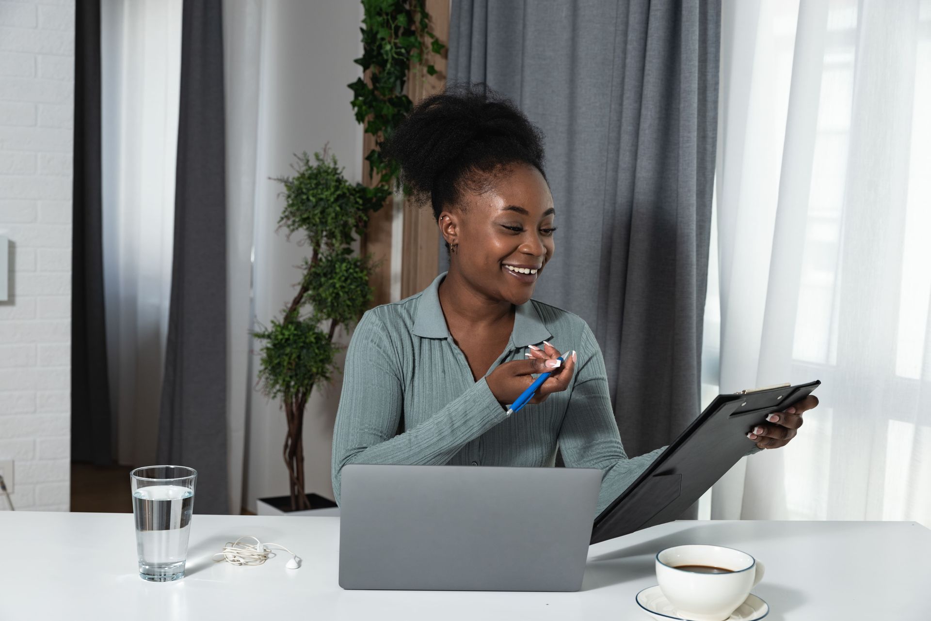 Woman on a video call at a desk, smiling with laptop, tablet, and earbuds in a bright room