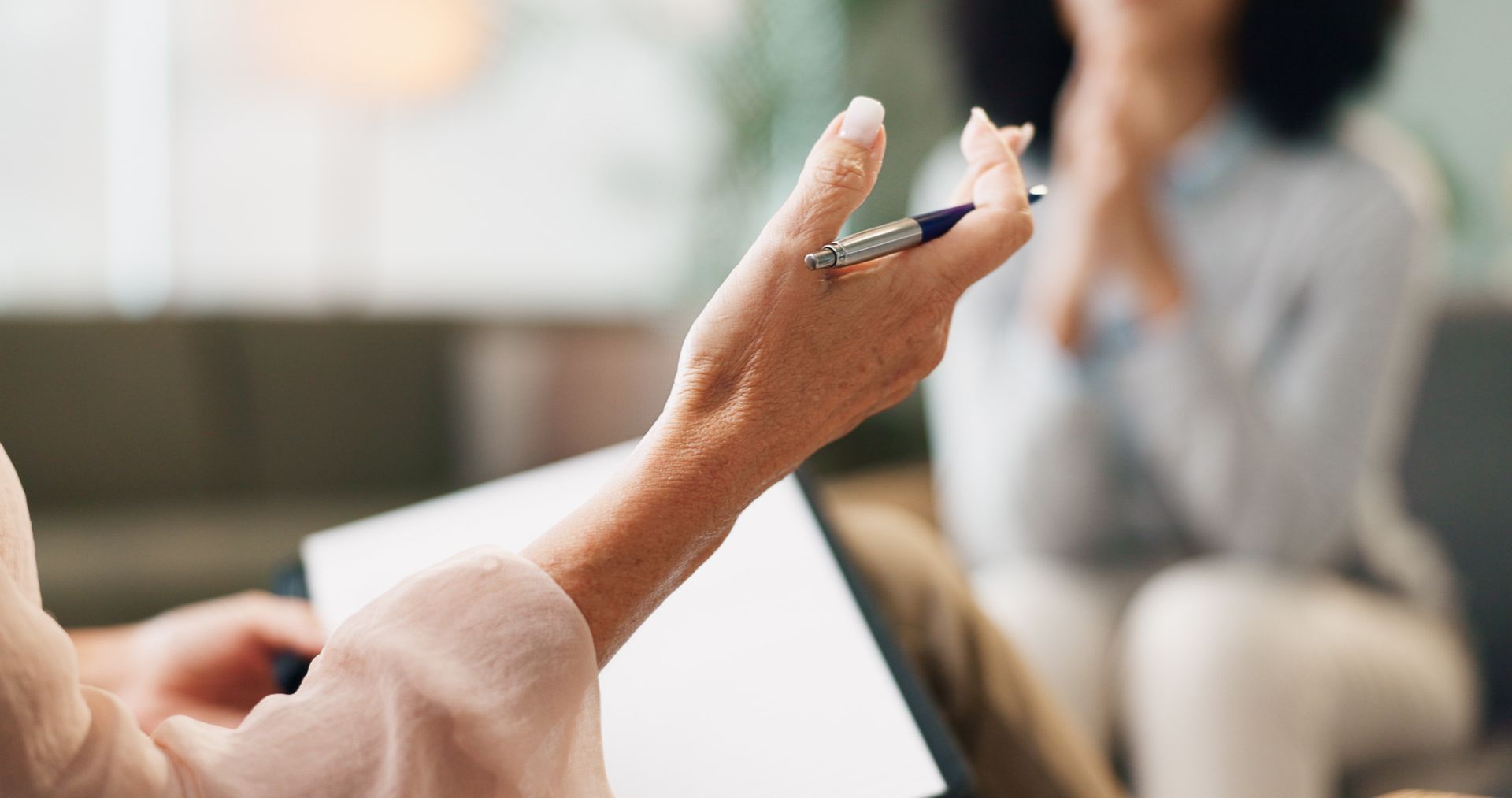 Hands gesturing in a blurred meeting room, with one person seated in the background holding a notebook.