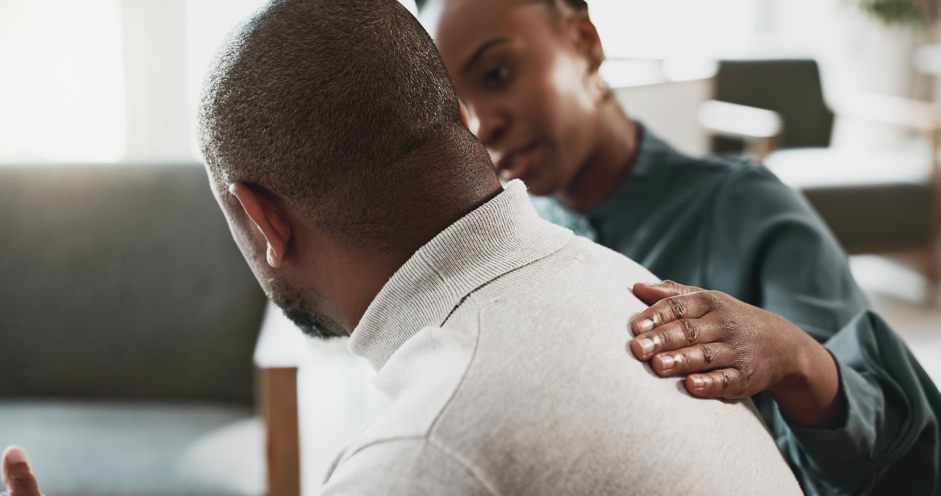 Person comforting another with a hand on their shoulder in a bright indoor room.