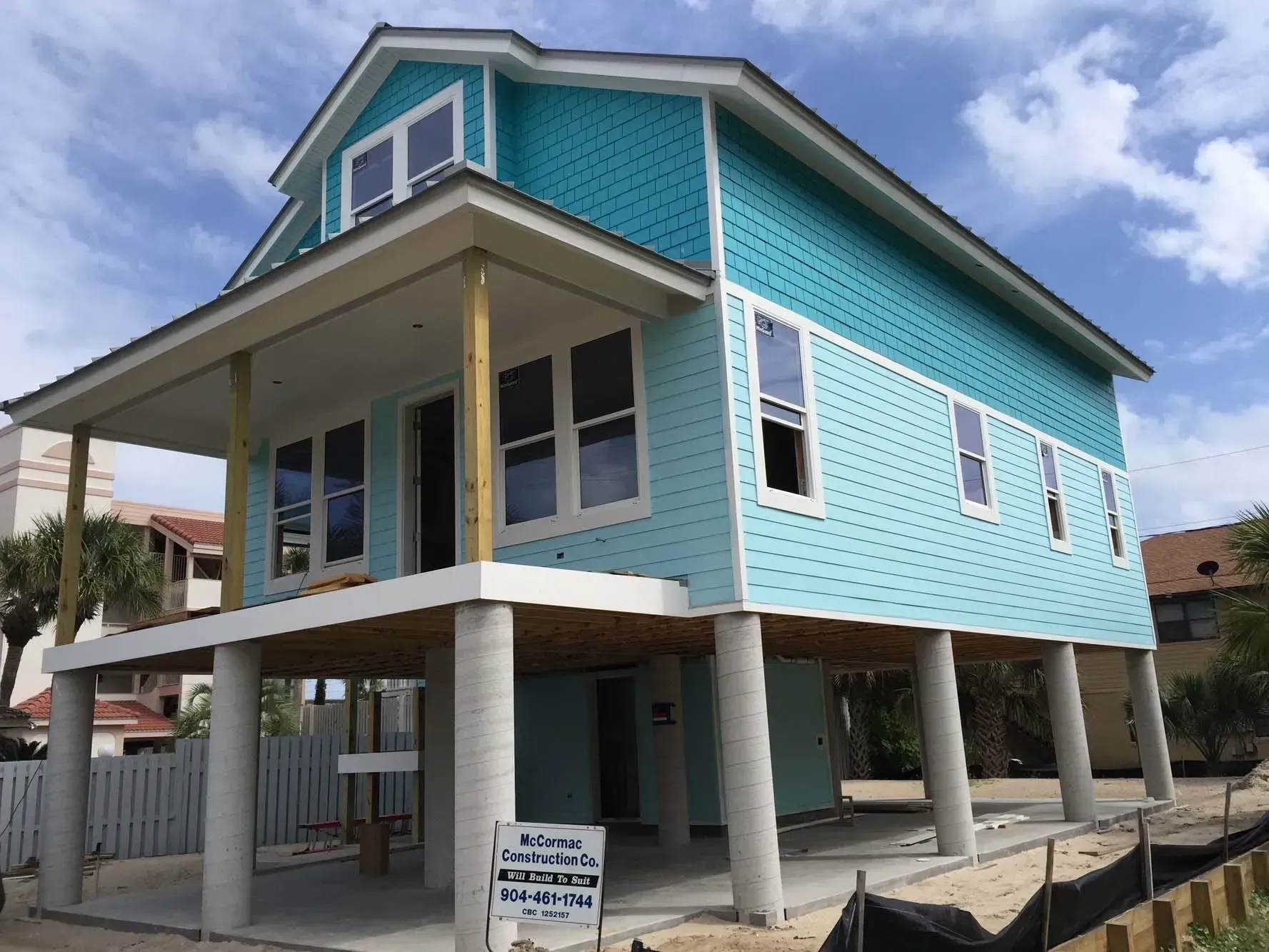 Elevated turquoise house with white trim on stilts, viewed from the front under a partly cloudy sky