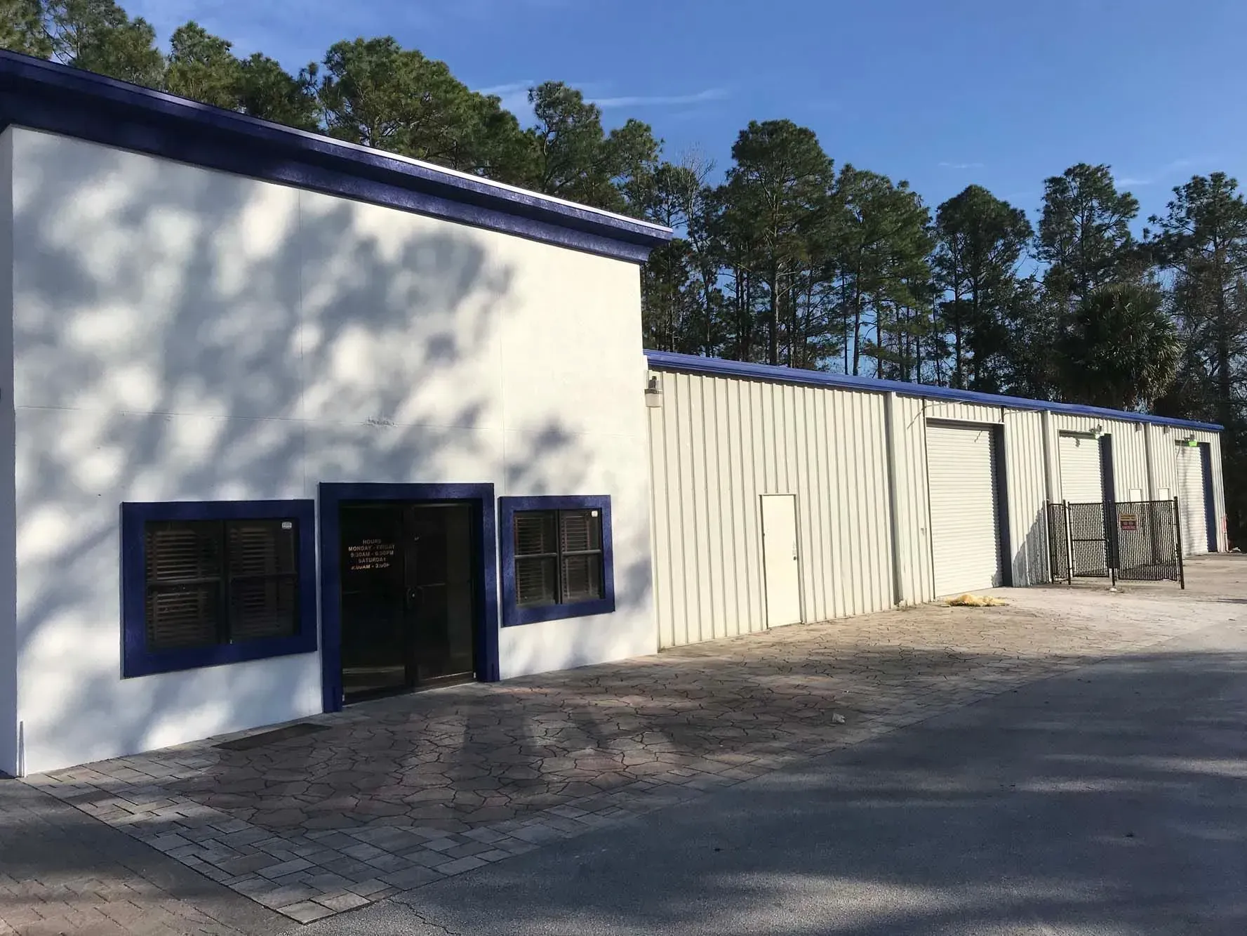 White industrial building with blue trim, black windows, and gravel parking lot under trees