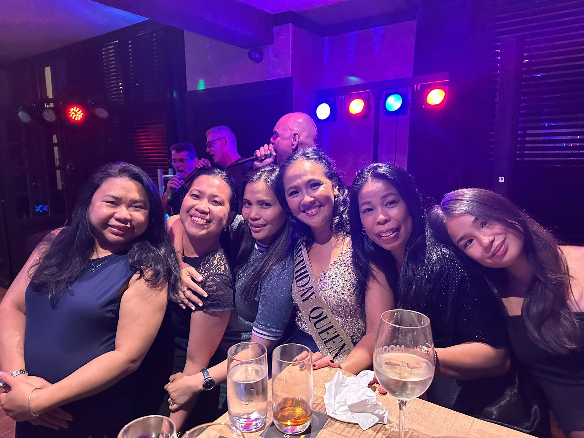 Group of women at a party, smiling, posing with drinks. Live band in the background. Dim lighting.
