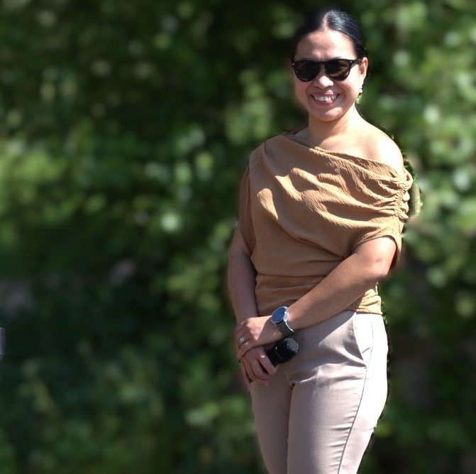 Woman in sunglasses smiles, wearing tan off-the-shoulder top and pants, standing outside.