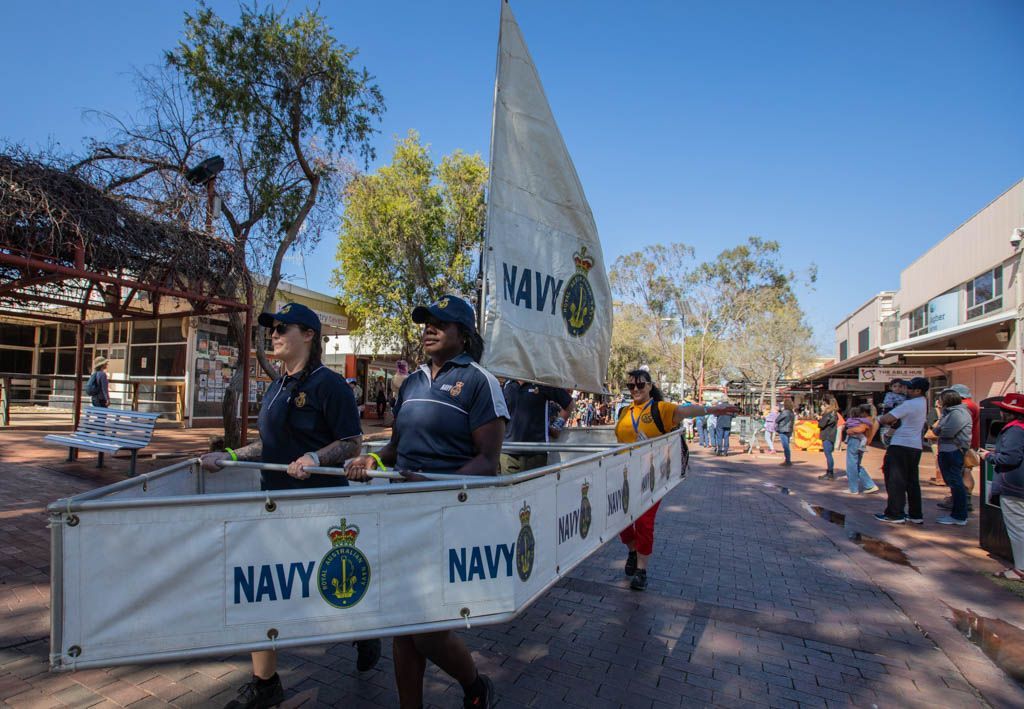 A group of people are walking down a street carrying a sailboat made out of a sign that says navy.
