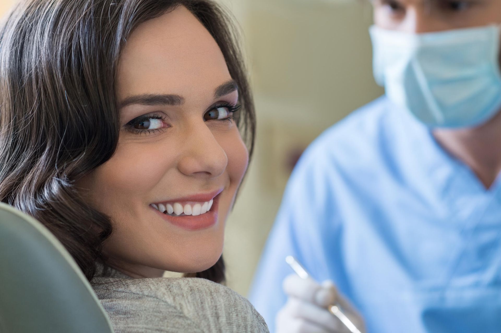 Woman smiles widely at the camera during a dental checkup; dentist in blue scrubs behind her.