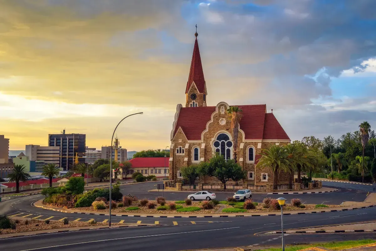 Historic church with red spire at a roundabout during sunset, with cars and city buildings in the background