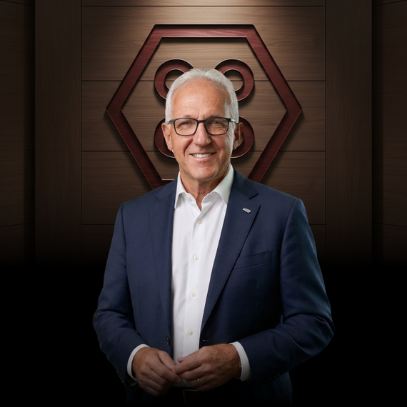 A smiling person in a blue blazer and white blouse stands before a dark wall with a hexagonal emblem.