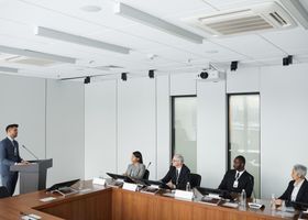 Business meeting in a bright conference room, with one speaker at a podium and several people seated at a long table.