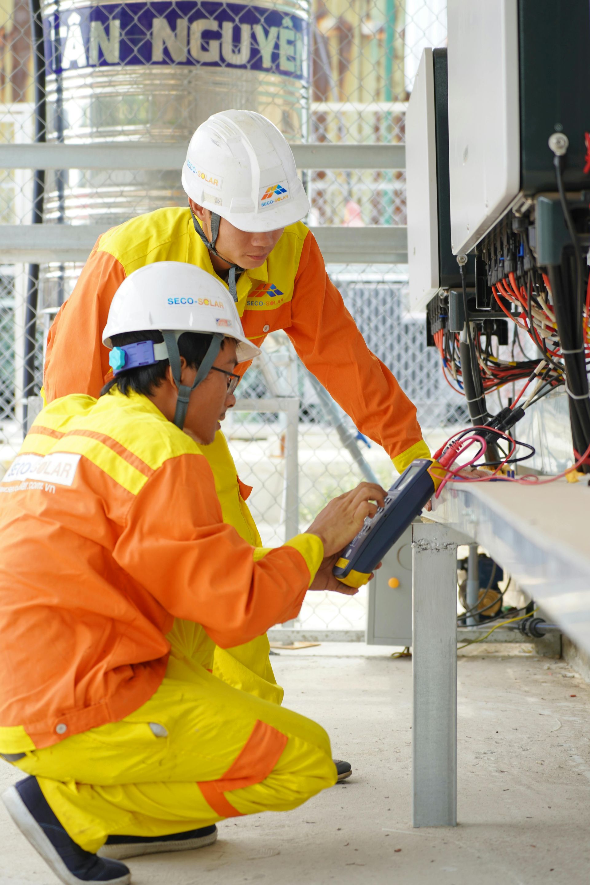 Two utility workers in orange and yellow safety suits and white helmets inspect electrical equipment outdoors.