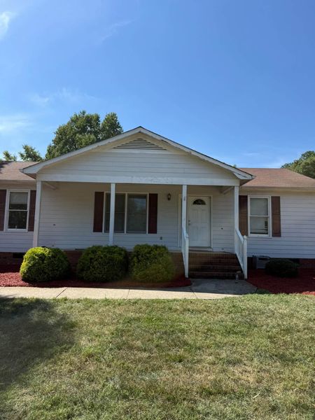 White single-story house with a front porch, red shutters, and a grassy lawn under a blue sky