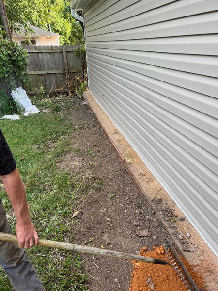 Person raking dirt along a house’s side yard beside gray vinyl siding and a wooden fence.