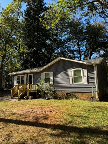 Gray house with white trim and front porch in a wooded yard, sunlit under tall trees