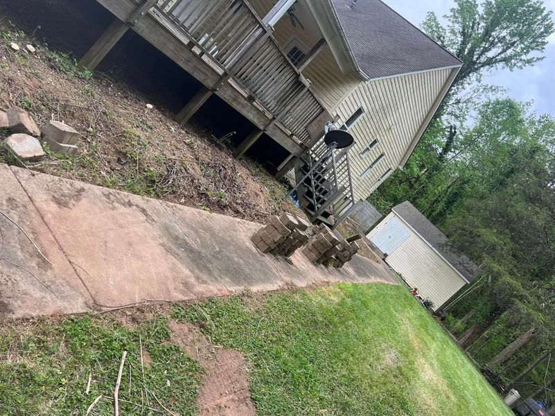 Tilted view of a house exterior with a concrete ramp, small shed, and trees in a grassy yard.
