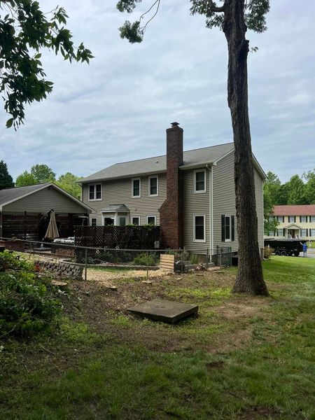 Backyard of a two-story house with a brick chimney, stone patio, and tall tree under cloudy skies