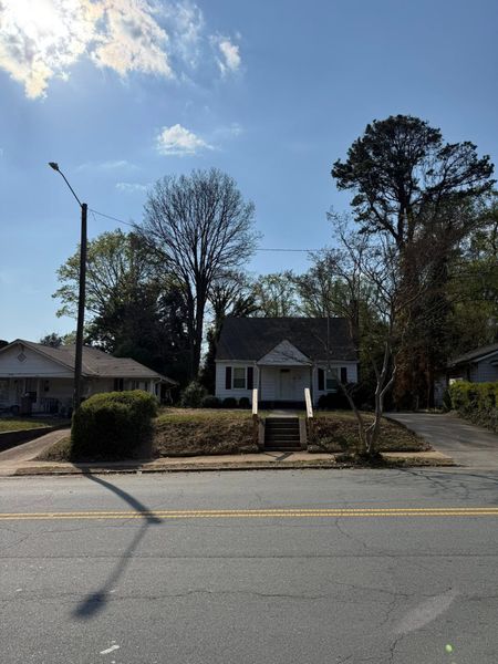Small white house with a front porch beside a road, framed by tall trees under a blue sky.