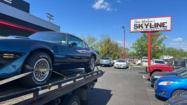 Classic dark blue car on a tow truck outside a Skyline dealership lot under a clear blue sky
