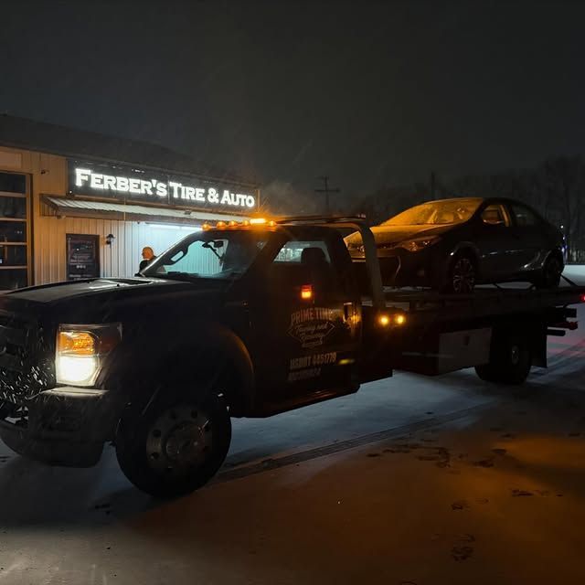 Tow truck hauling a dark sedan at night outside Ferber’s Tire & Auto, with lights glowing in snow.