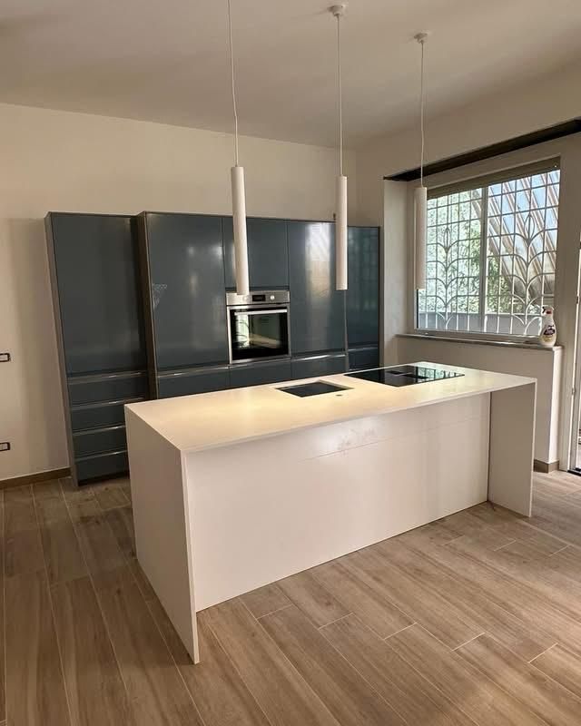 Cucina moderna con isola bianca, mobili scuri, lampade a sospensione e una grande finestra.
