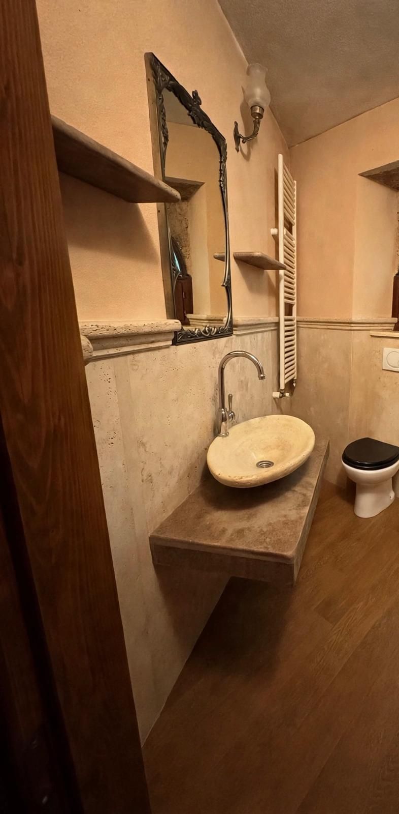 Piccolo bagno con lavabo a parete, specchio, mensole, WC e pareti rivestite in legno.