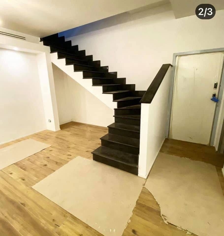 Stanza vuota con una scala nera, pareti bianche e pavimento in legno grezzo e compensato.