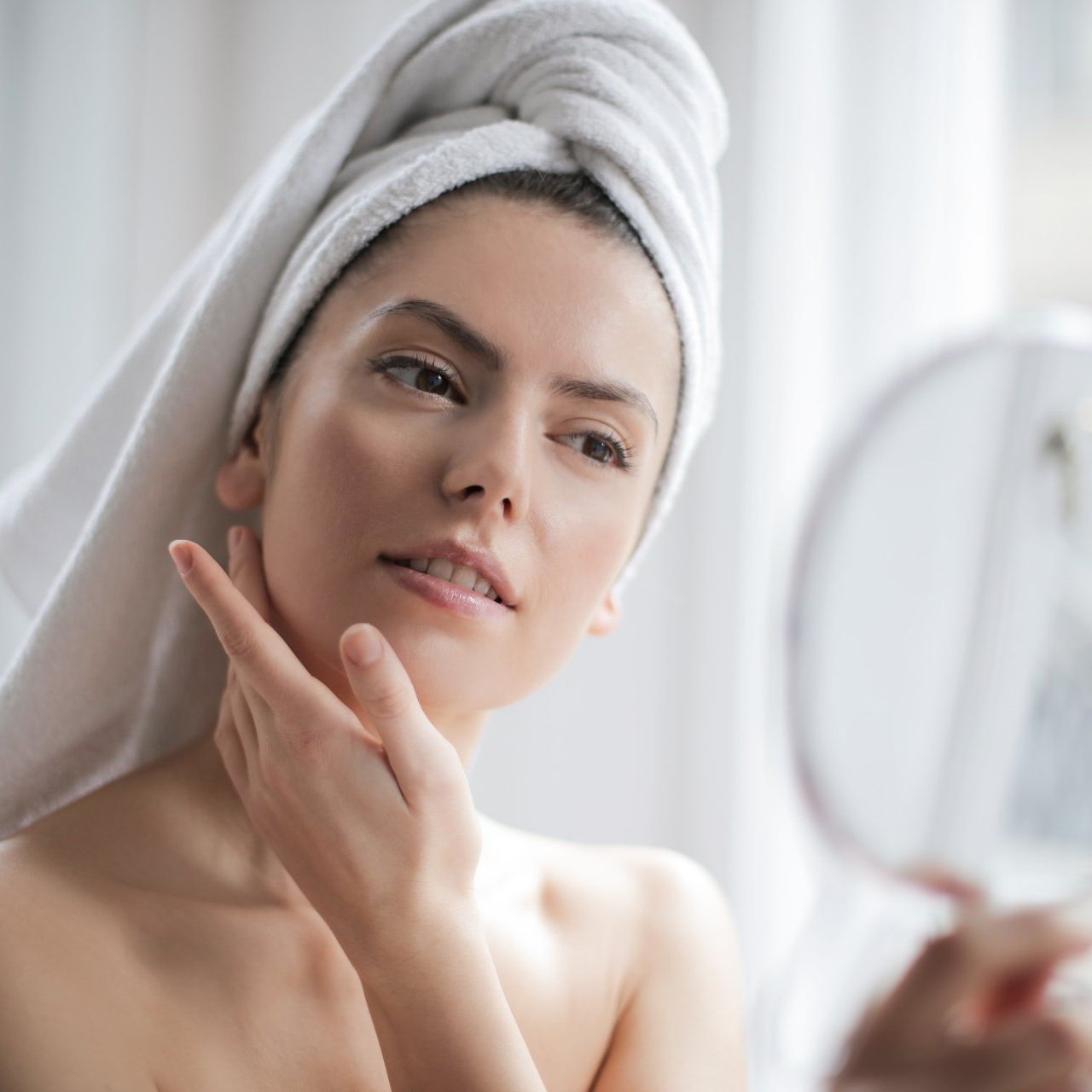 A woman with a towel wrapped around her head is looking at her face in a mirror.