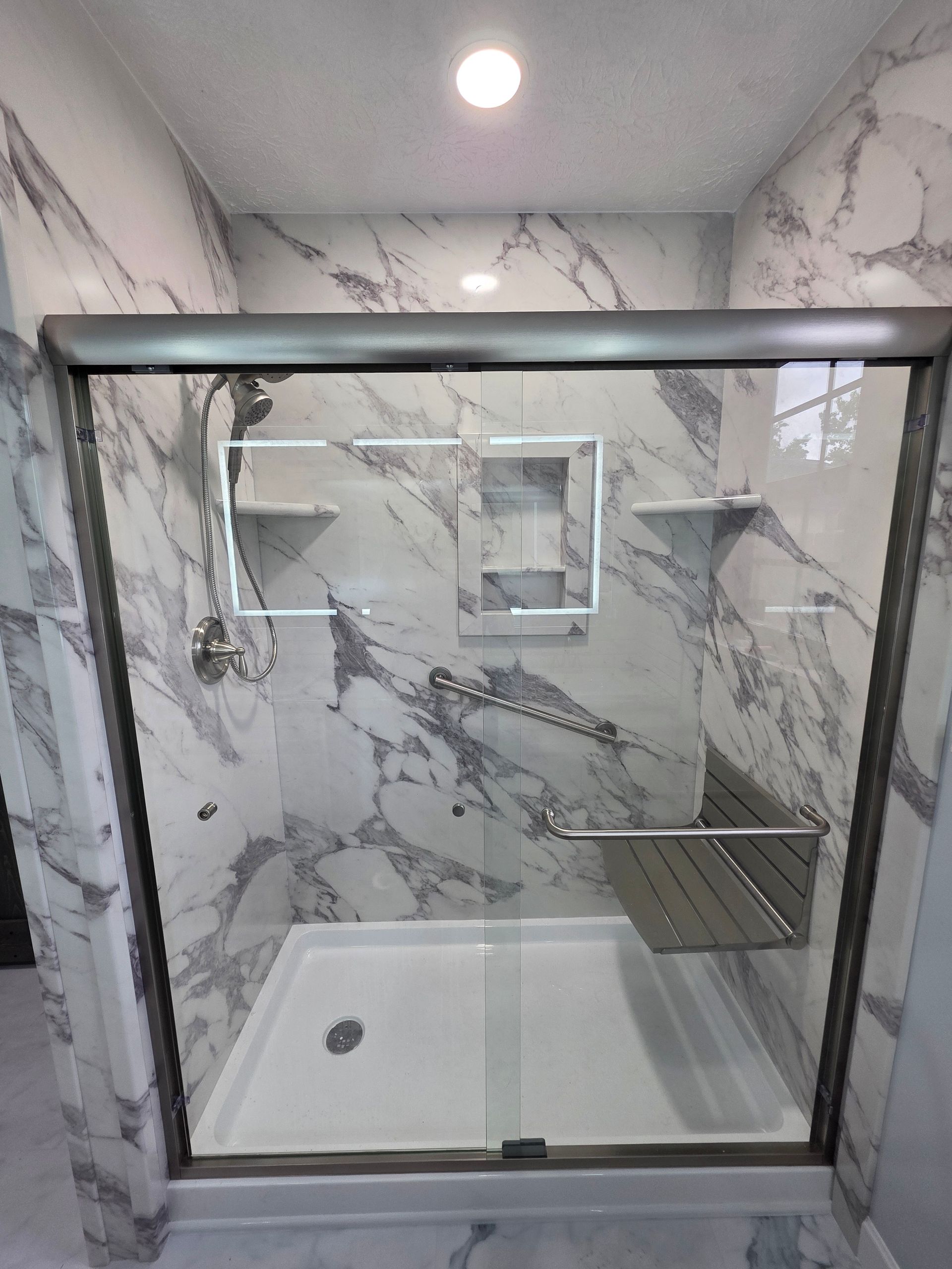 Walk-in shower with marble-look tile, glass door, shelf, and overhead light.