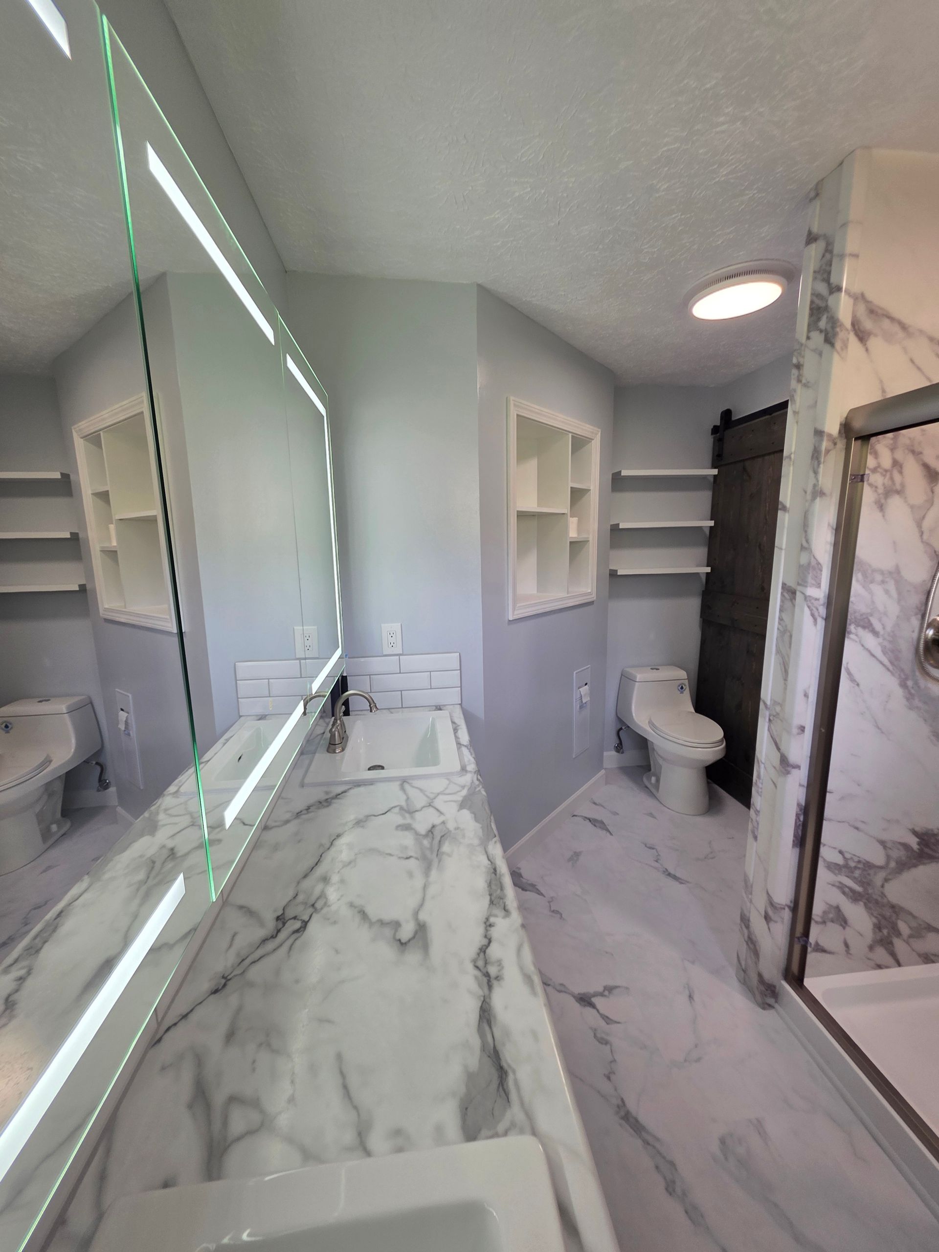 Modern bathroom with marble vanity, large mirror, white shelves, toilet, and glass shower enclosure