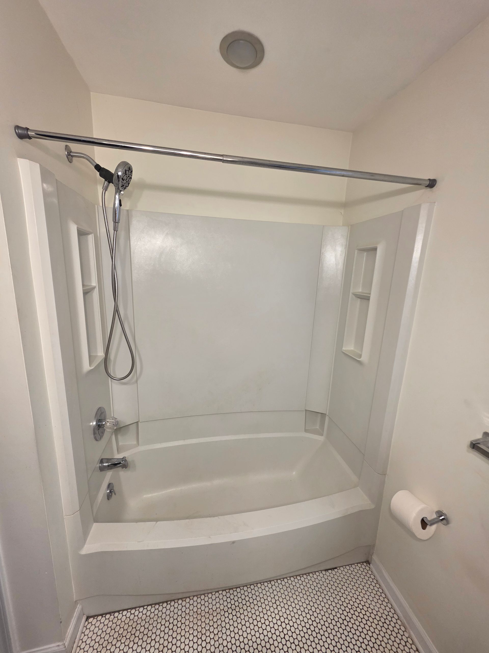 White bathroom with a tub, shower curtain rod, handheld showerhead, and patterned floor mat.