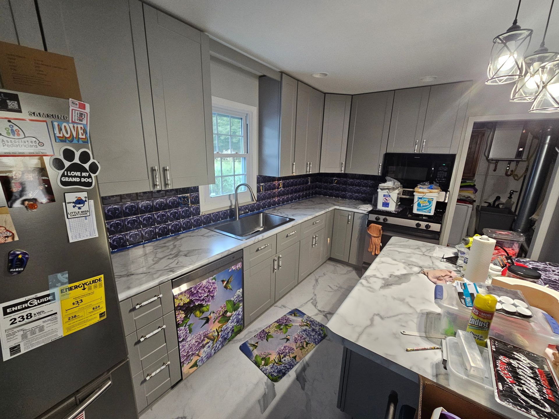 Gray kitchen with white countertops and purple tile backsplash. Refrigerator, cabinets, and sink are visible.