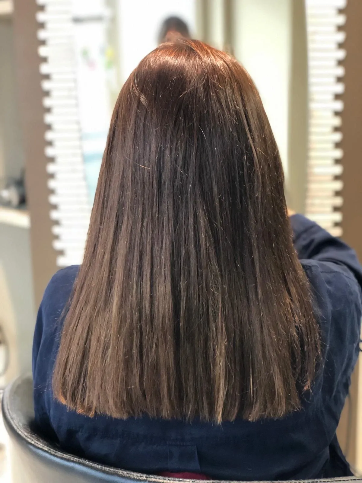 Eine Person mit glattem, braunem Haar, von hinten gesehen, sitzt in einem Friseursalon auf einem Friseurstuhl.