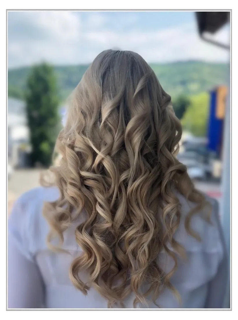 Welliges blondes Haar fällt kaskadenartig den Rücken hinab, im Hintergrund ist die Natur verschwommene Atmosphäre zu sehen.