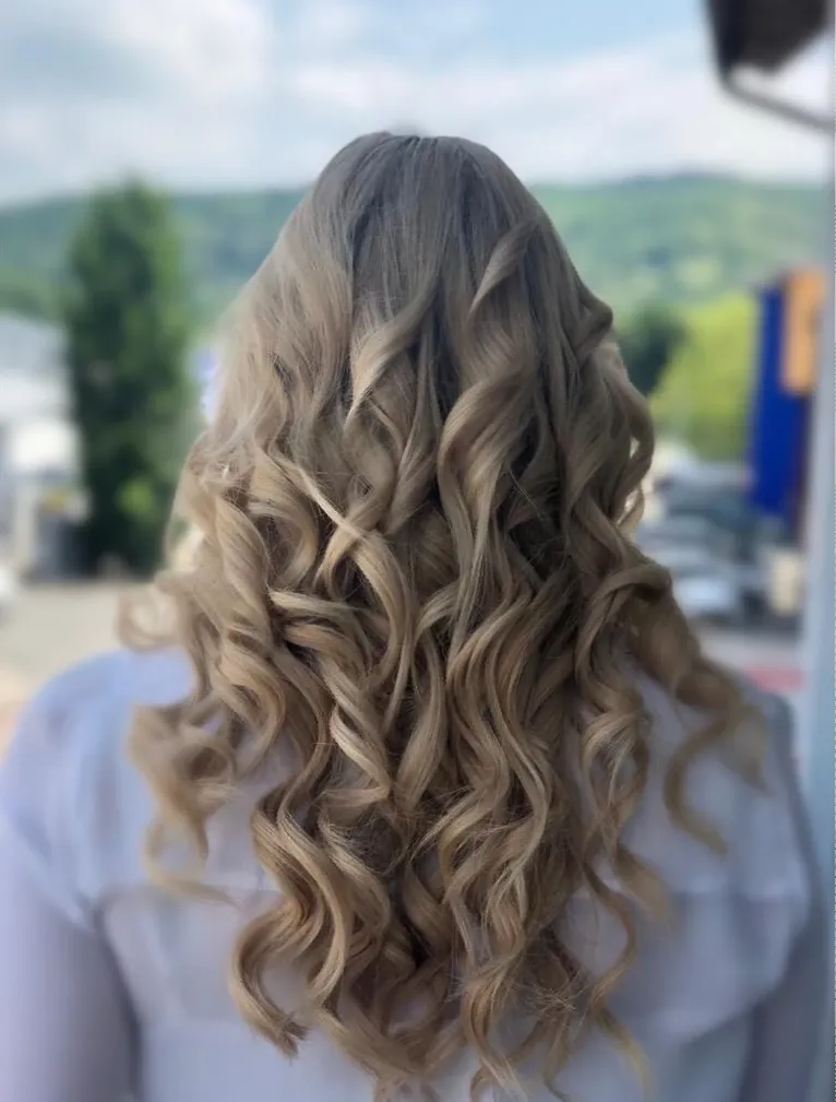 Welliges blondes Haar fällt vor einem verschwommenen Hintergrund im Freien den Rücken hinab.