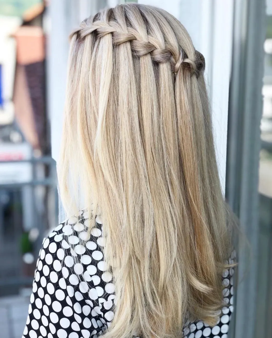 Rückansicht von langem blondem Haar mit einem geflochtenen Haarkranz, dazu ein schwarz-weiß gepunktetes Oberteil.